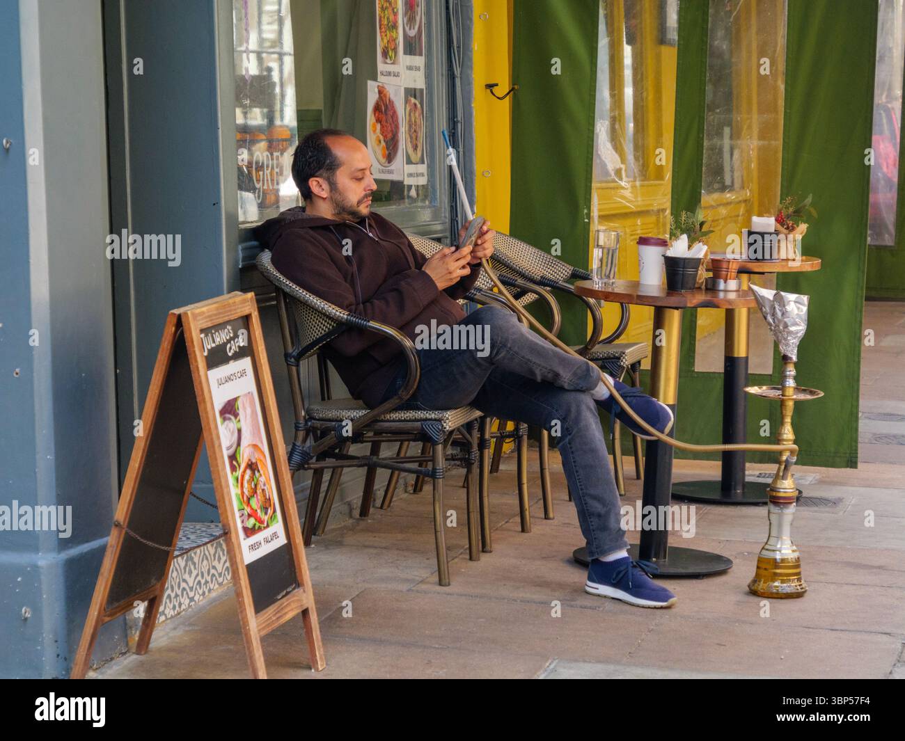 Un uomo si diverte a fare una seduta di narghilè mentre usa il suo smartphone fuori dal Juliano's Café di Mayfair, Londra, catturando un mix di vita digitale moderna e cultura dei caffè mediorientali in uno dei quartieri più esclusivi della città Foto Stock