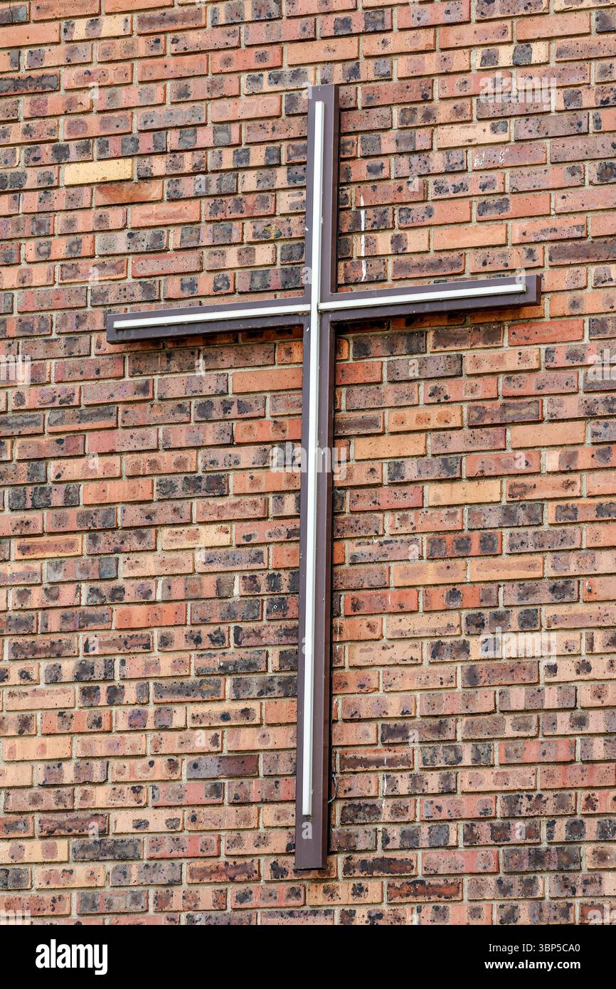 Una croce cristiana metallica montata su un muro di mattoni rossi, simbolo di fede e spiritualità all'esterno di una chiesa Foto Stock