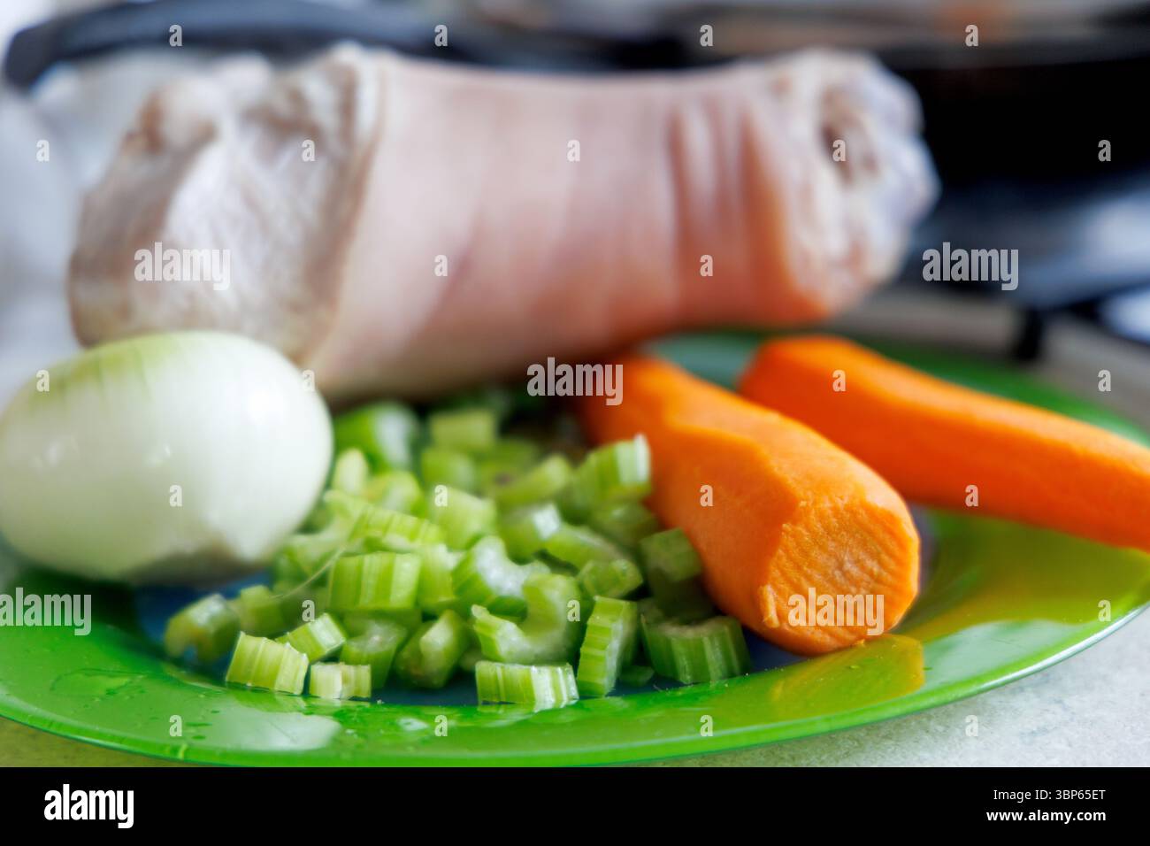 Uno stinco di maiale sbucciato è visibile su un piatto verde, accanto ad esso c'è mezza cipolla, gambi di sedano tritati e due carote. Foto Stock