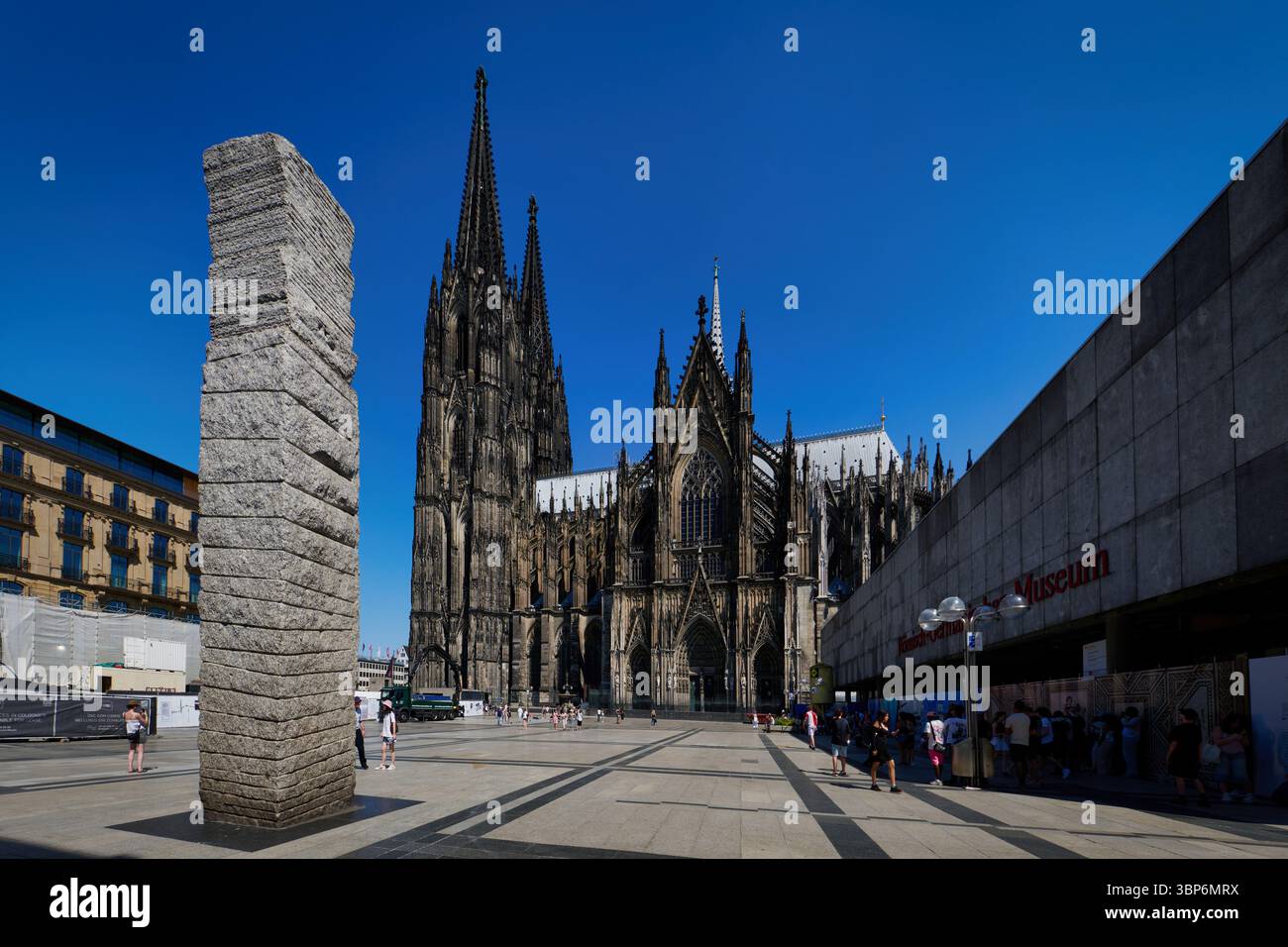 Colonia, Germania 01 luglio 2025: Vista sulla vivace Roncalliplatz verso la cattedrale di Colonia, incorniciata dalla colonna della Pace e dal Museo romano-germanico Foto Stock