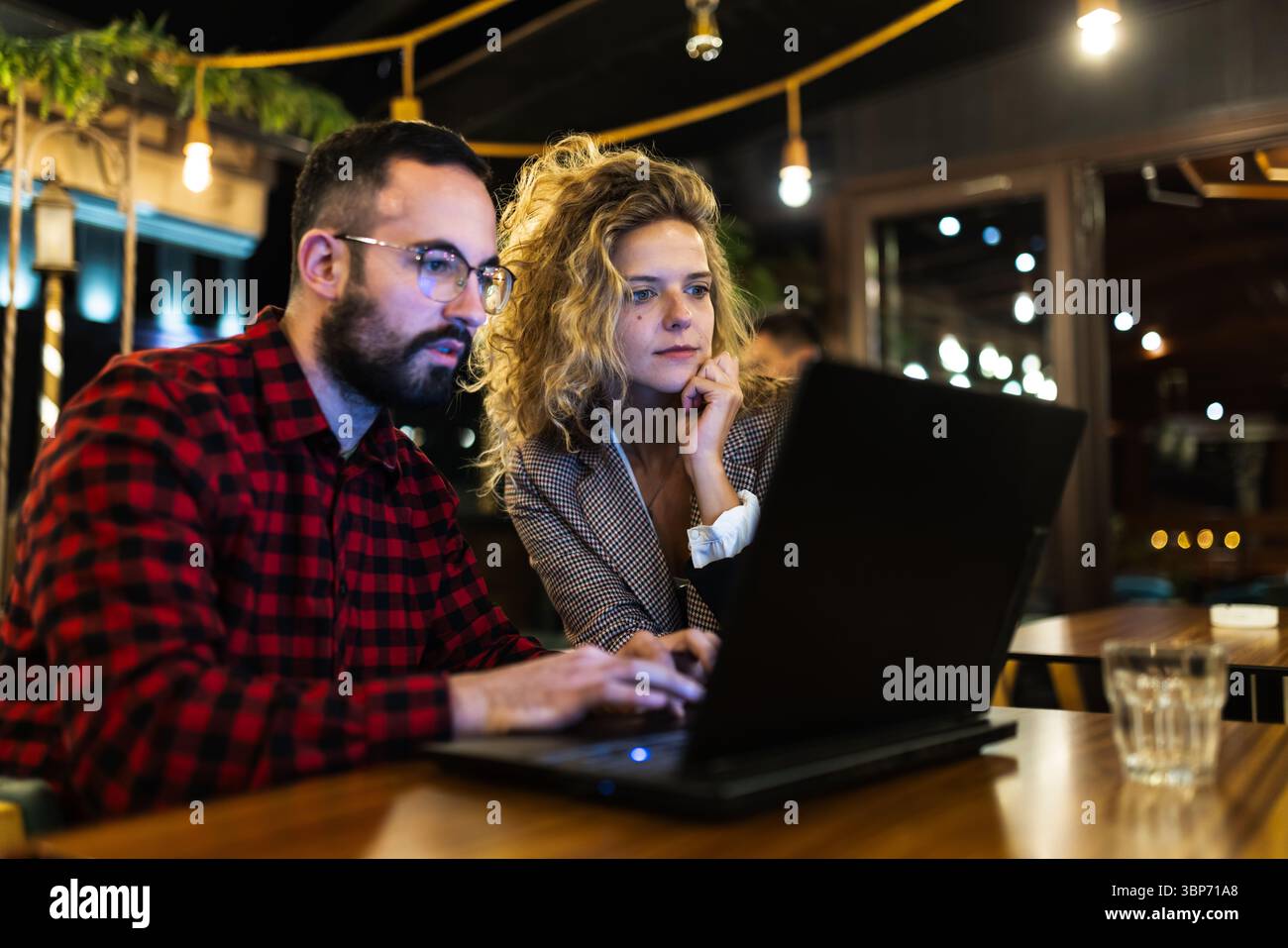 Due colleghi si concentrano intensamente su un notebook in uno spazio di lavoro accogliente ed elegante, evidenziando il lavoro di squadra, la concentrazione e la collaborazione durante la serata Foto Stock