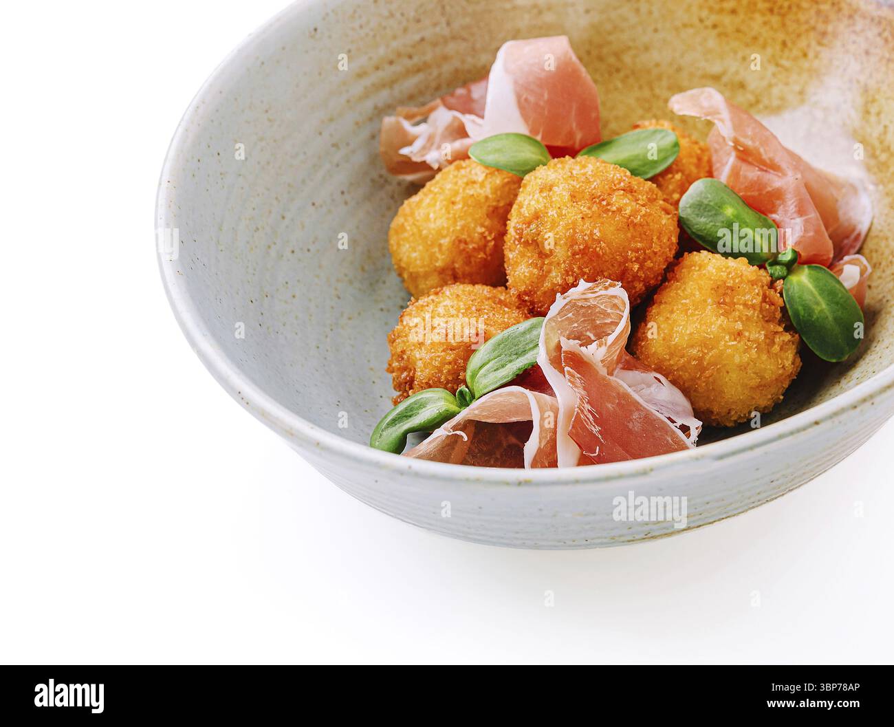 Cibo: Una ciotola presenta polpette fritte dorate croccanti accompagnate da sottili fette di prosciutto e foglie verdi fresche. Il piatto è presentato elegantemente, spettacolo Foto Stock