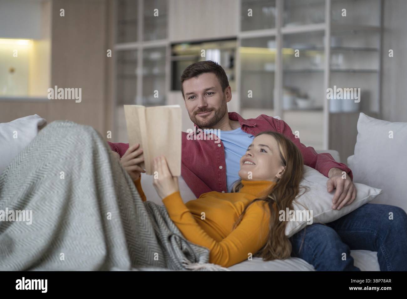Marito soddisfatto, moglie sul divano in soggiorno. Una donna allegra giace sul cuscino in braccio all'uomo, tenendo in mano un libro, leggendo ad alta voce con un sorriso. Felice ragazzo calmo ad ascoltare Foto Stock