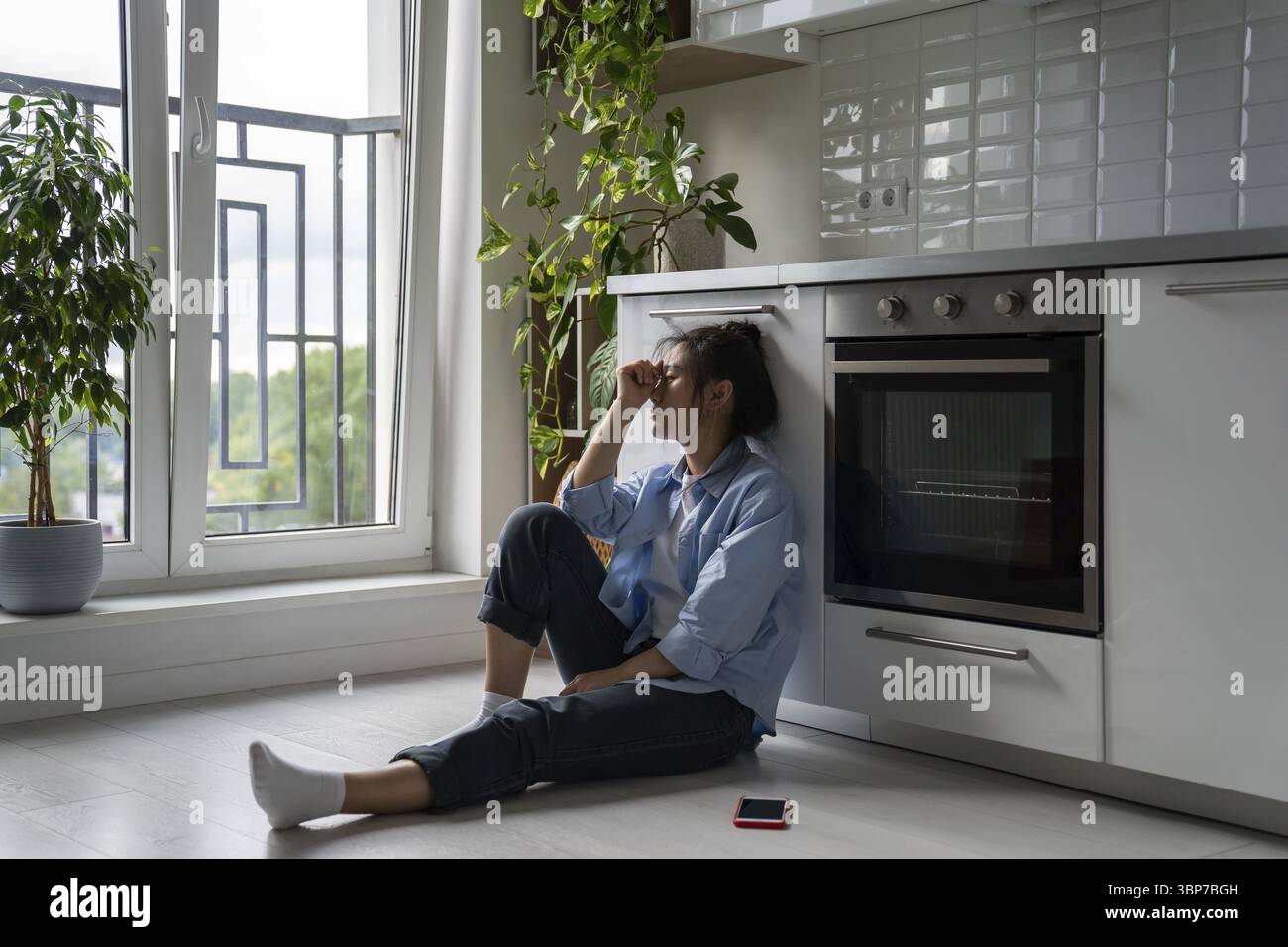 Infelice ragazza asiatica seduta sul pavimento in cucina si sente depressa dopo l'uso dei social media, viene ignorata dal fidanzato, affrontando il rifiuto in amore. Foto Stock