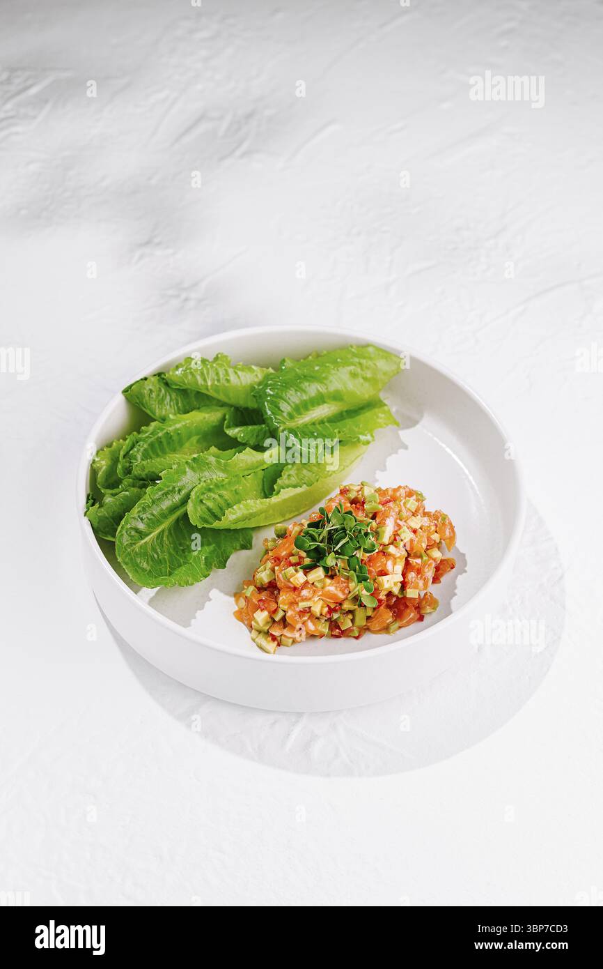 Cibo, tartare di salmone servita con avocado, erbe aromatiche e lattuga su piatto bianco su sfondo bianco Foto Stock