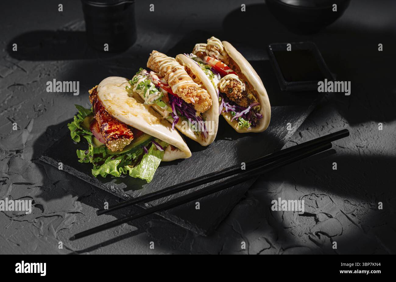 Cibo, tre panini bao ripieni di pollo croccante, lattuga fresca e verdure colorate riposano su un piatto di ardesia scuro, per un'appetitosa cucina Foto Stock