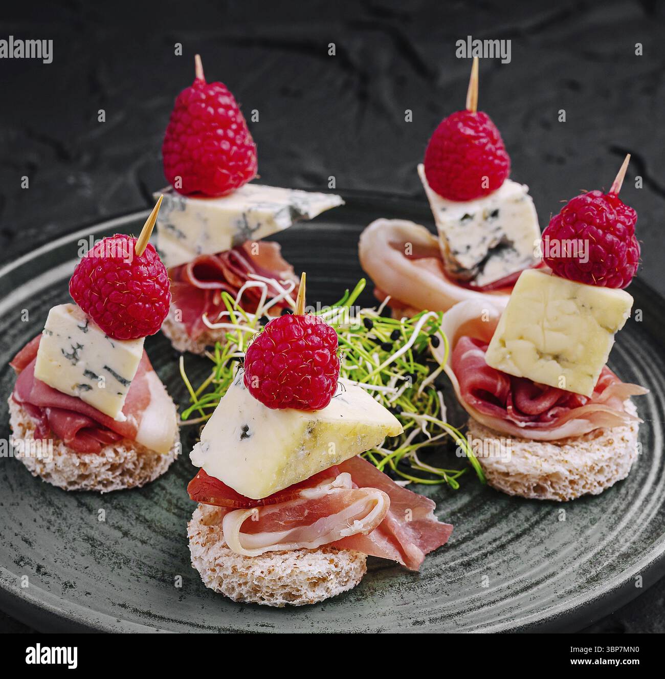 Cibo, deliziosi piccoli bocconcini con strati di formaggio, prosciutto e lamponi freschi su un piatto scuro. Perfetto per riunioni o cene di lusso Foto Stock