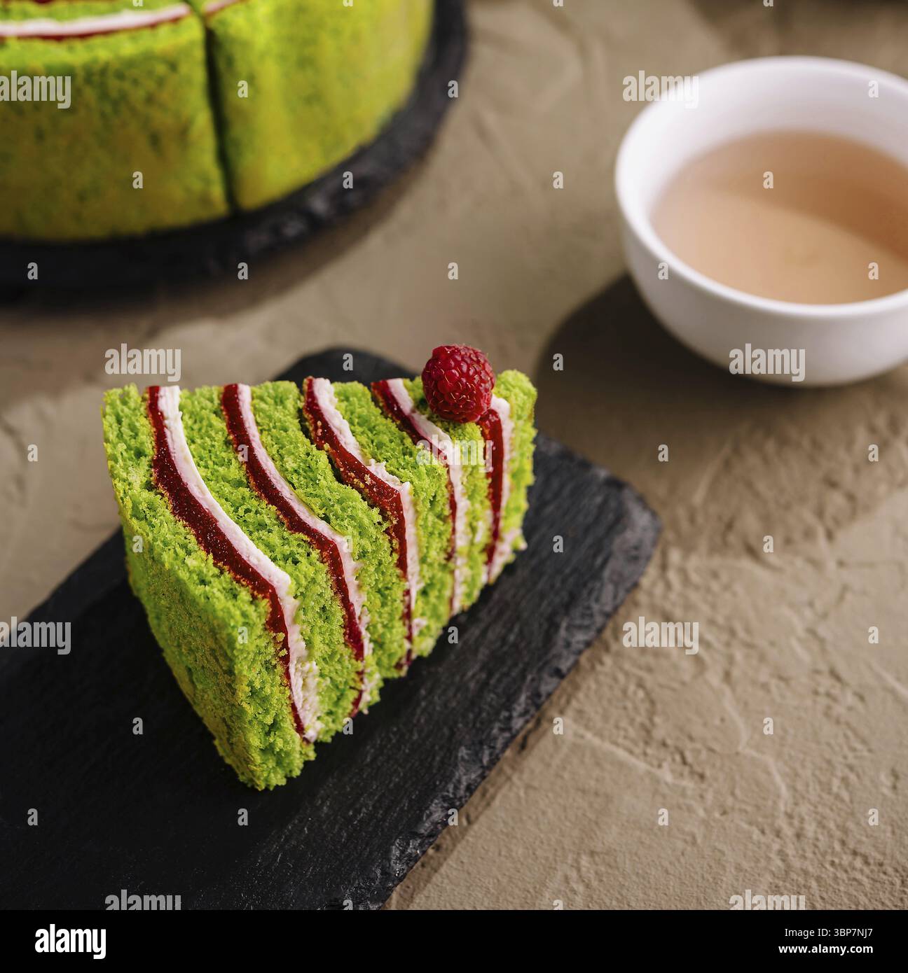 Cibo, fetta di vivace torta matcha di tè verde, decorata con lamponi, accanto a una torta completa e una tazza di tè Foto Stock
