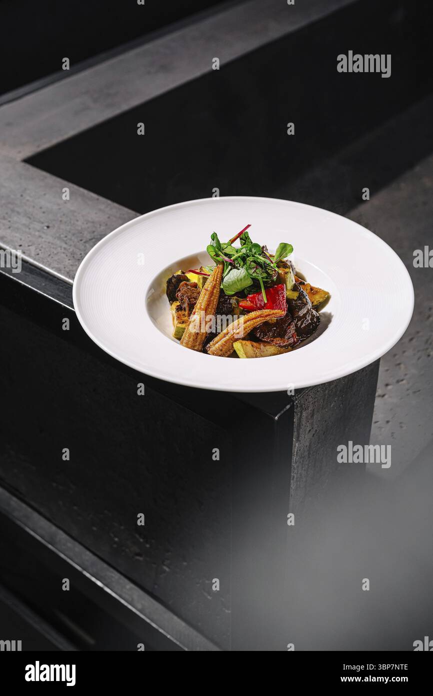 Cibo, Un piatto ben placcato presenta verdure grigliate insieme a carne tenera, guarnite con erbe fresche. L'ambiente minimalista aggiunge raffinatezza Foto Stock