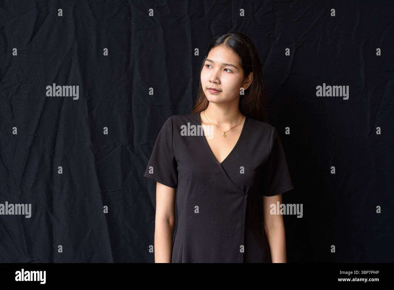 Giovane donna asiatica in abito nero che posa contro la tenda nera in luce naturale Foto Stock