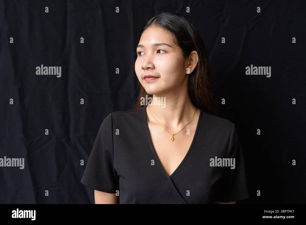 Giovane donna asiatica in abito nero che posa contro la tenda nera in luce naturale Foto Stock