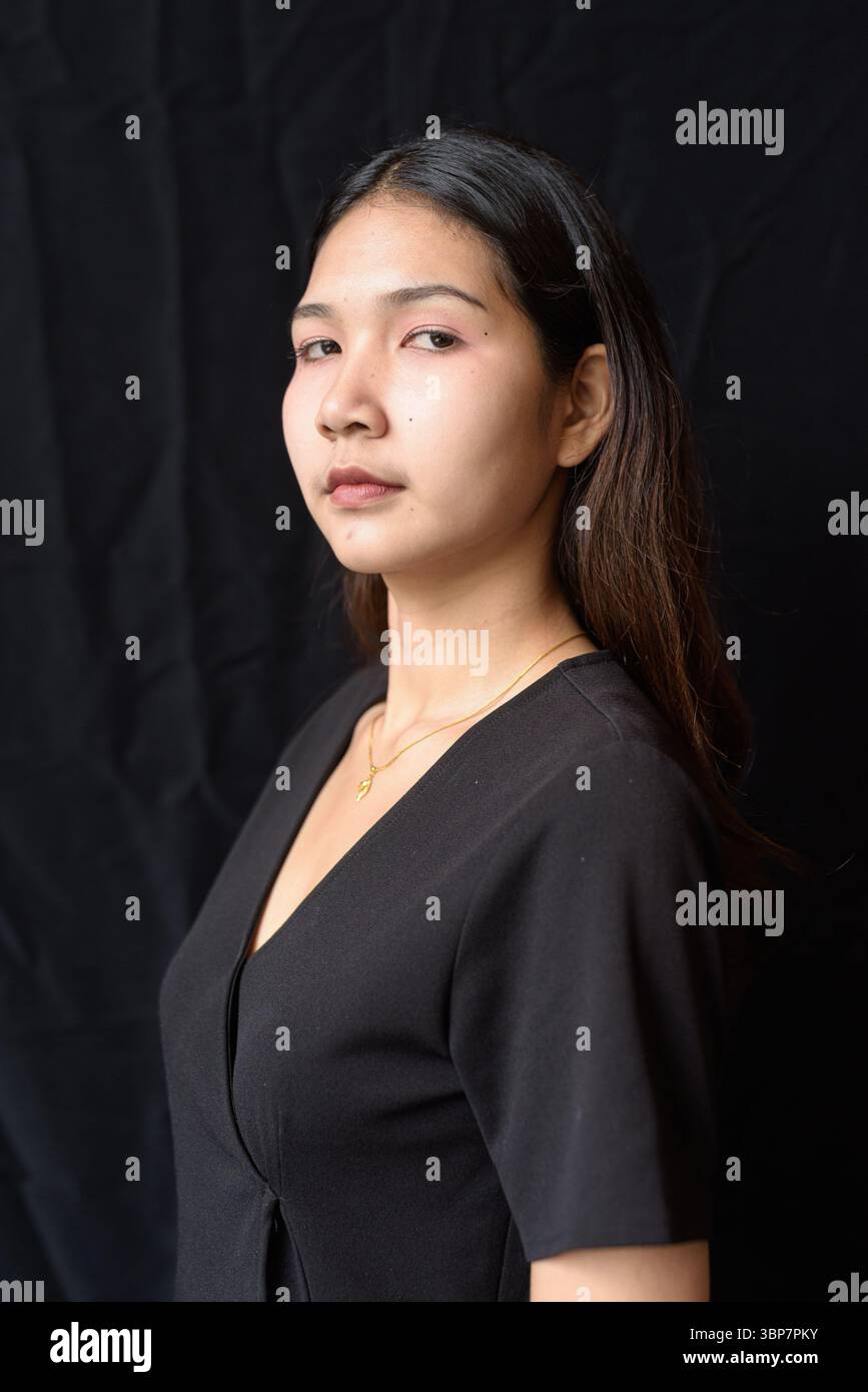 Giovane donna asiatica in abito nero che posa contro la tenda nera in luce naturale Foto Stock
