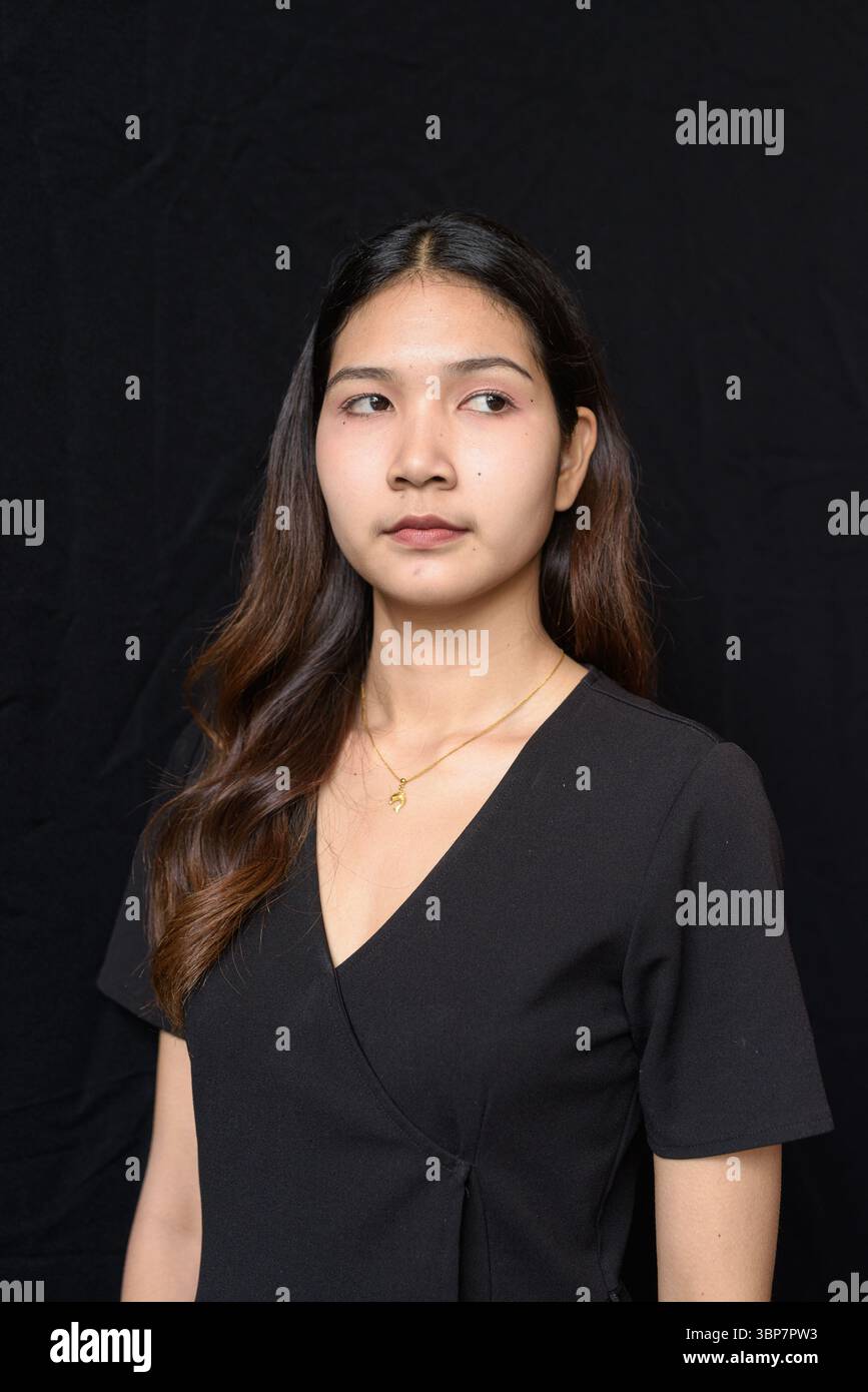 Giovane donna asiatica in abito nero che posa contro la tenda nera in luce naturale Foto Stock