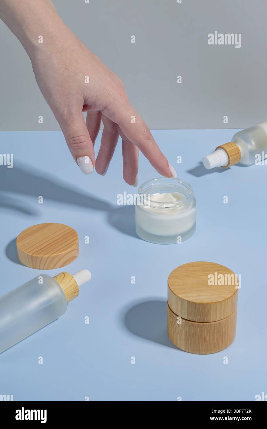 Donna che applica crema sulla mano su sfondo blu con prodotti cosmetici e ombre dure. Concetto di trattamento per la cura della pelle Foto Stock