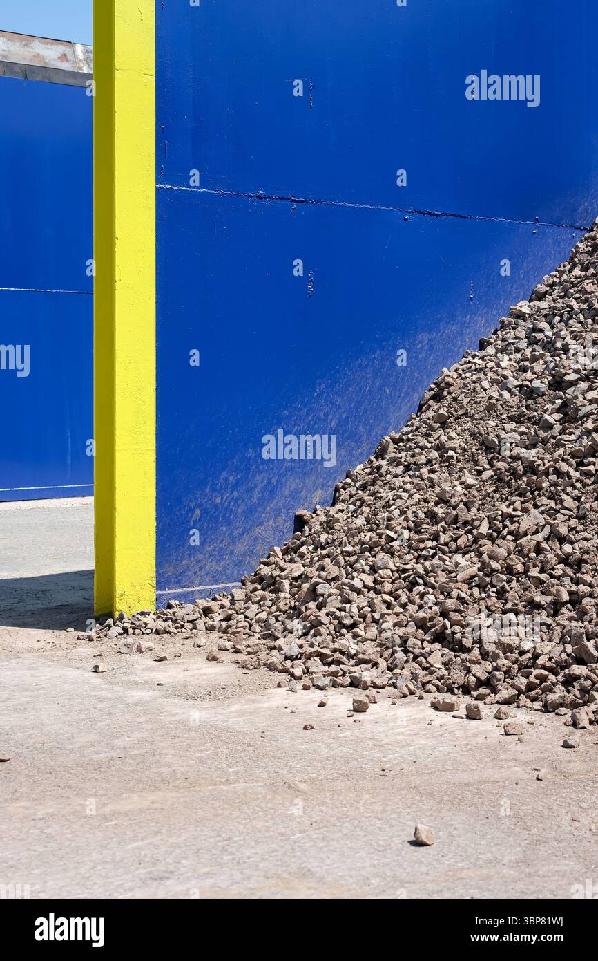 Scena industriale con grande palo di pietra frantumata o ghiaia. Parete metallica blu o barriera con montante giallo. Materiale da costruzione. Foto Stock