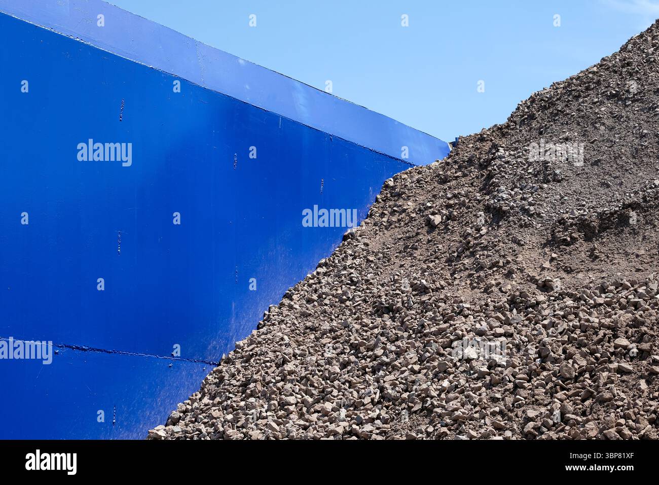 Grande pila di pietra schiacciata o ghiaia. Parete o barriera metallica blu. Materiale da costruzione in un cantiere industriale o in una base di costruzione. Copia spazio. Foto Stock