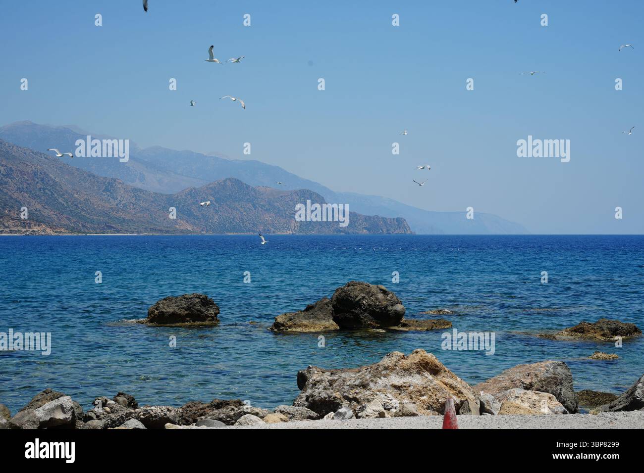 I gabbiani volano graziosamente sopra le limpide acque del Mediterraneo, con formazioni rocciose in primo piano e montagne lontane all'orizzonte. Cieli blu Foto Stock