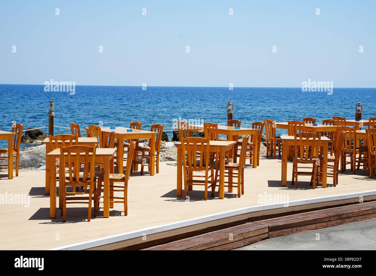 Terrazza vuota all'aperto con tavoli e sedie in legno che si affaccia su un oceano calmo. L'allestimento è perfetto per pranzare all'aperto, offrendo un ambiente sereno Foto Stock