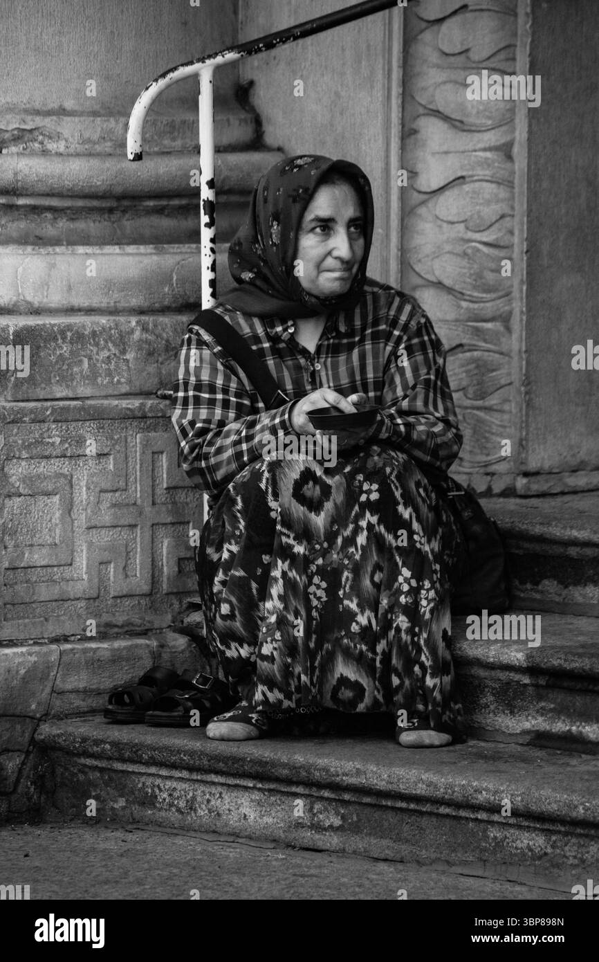 Anziana zingara che implorava da sola in strada seduta tranquillamente in abiti a fantasia concetto di povertà catturato in ambiente urbano emozioni umane grezze Foto Stock