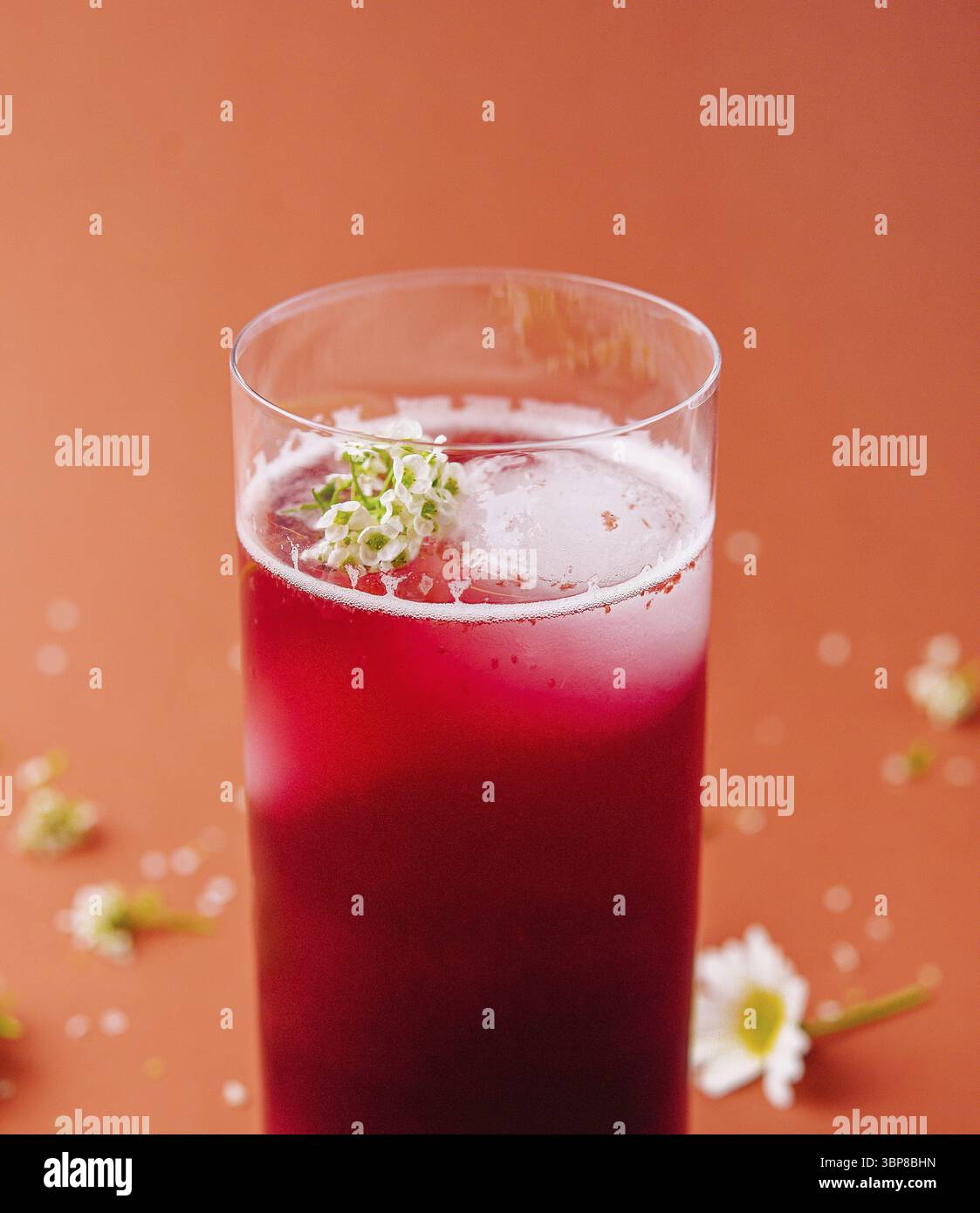 Cibo, vivace frullato di frutti di bosco rossi in un alto bicchiere, decorato con piccoli fiori bianchi su uno sfondo arancione Foto Stock