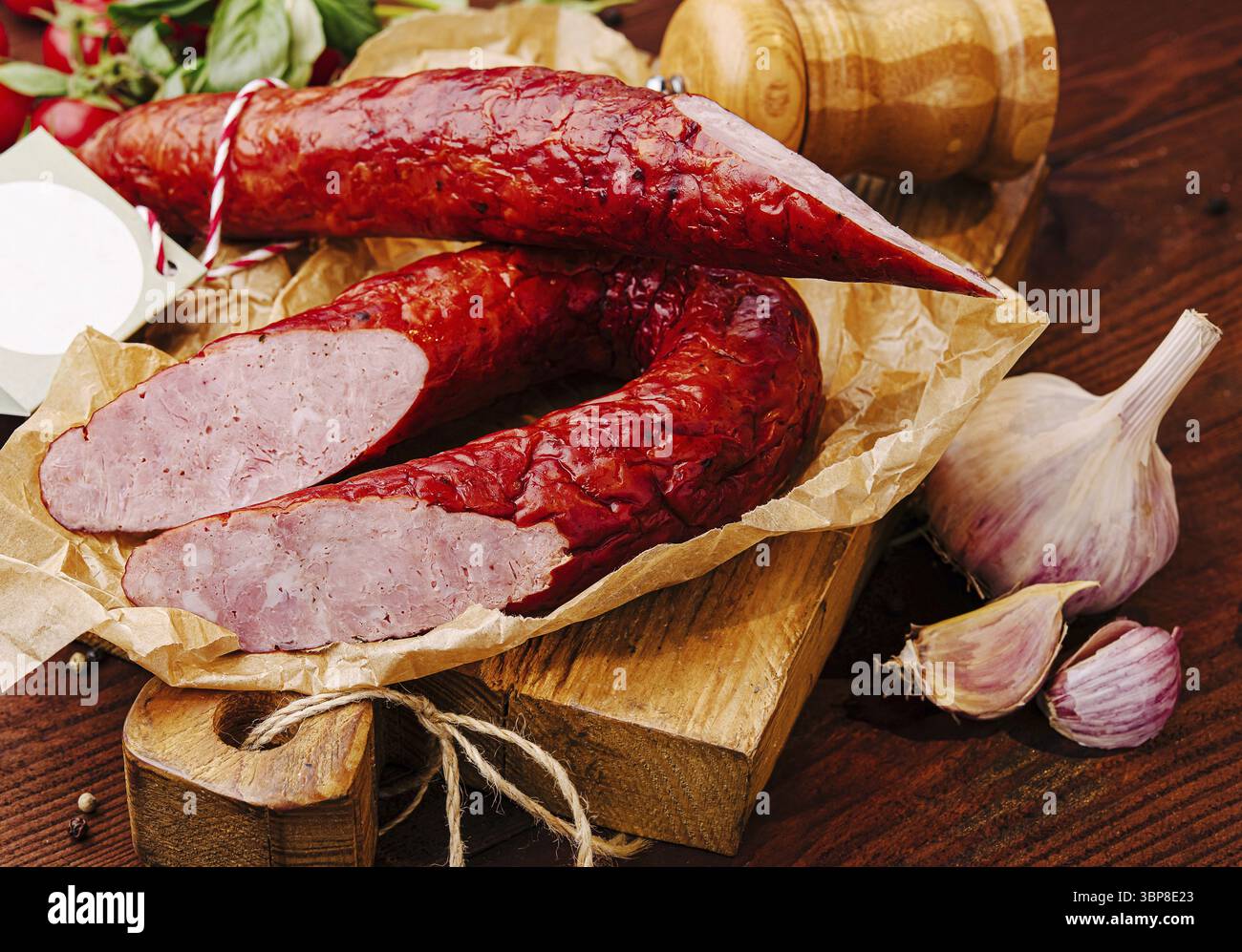 Cibo, salsiccia affumicata appena tagliata si trova su un tavolo di legno circondato da aglio ed erbe aromatiche. I ricchi colori e le texture ne esaltano l'appetitoso fascino Foto Stock