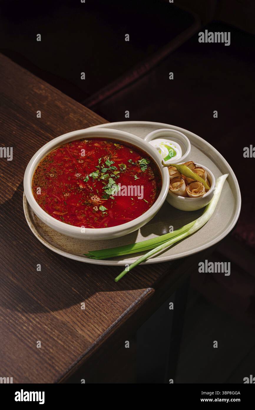 Cibo, Una ciotola di zuppa rossa ricca e piccante è posta su un tavolo di legno, guarnita con erbe fresche. Ad accompagnare la zuppa ci sono cipolle verdi e un piccolo piatto Foto Stock