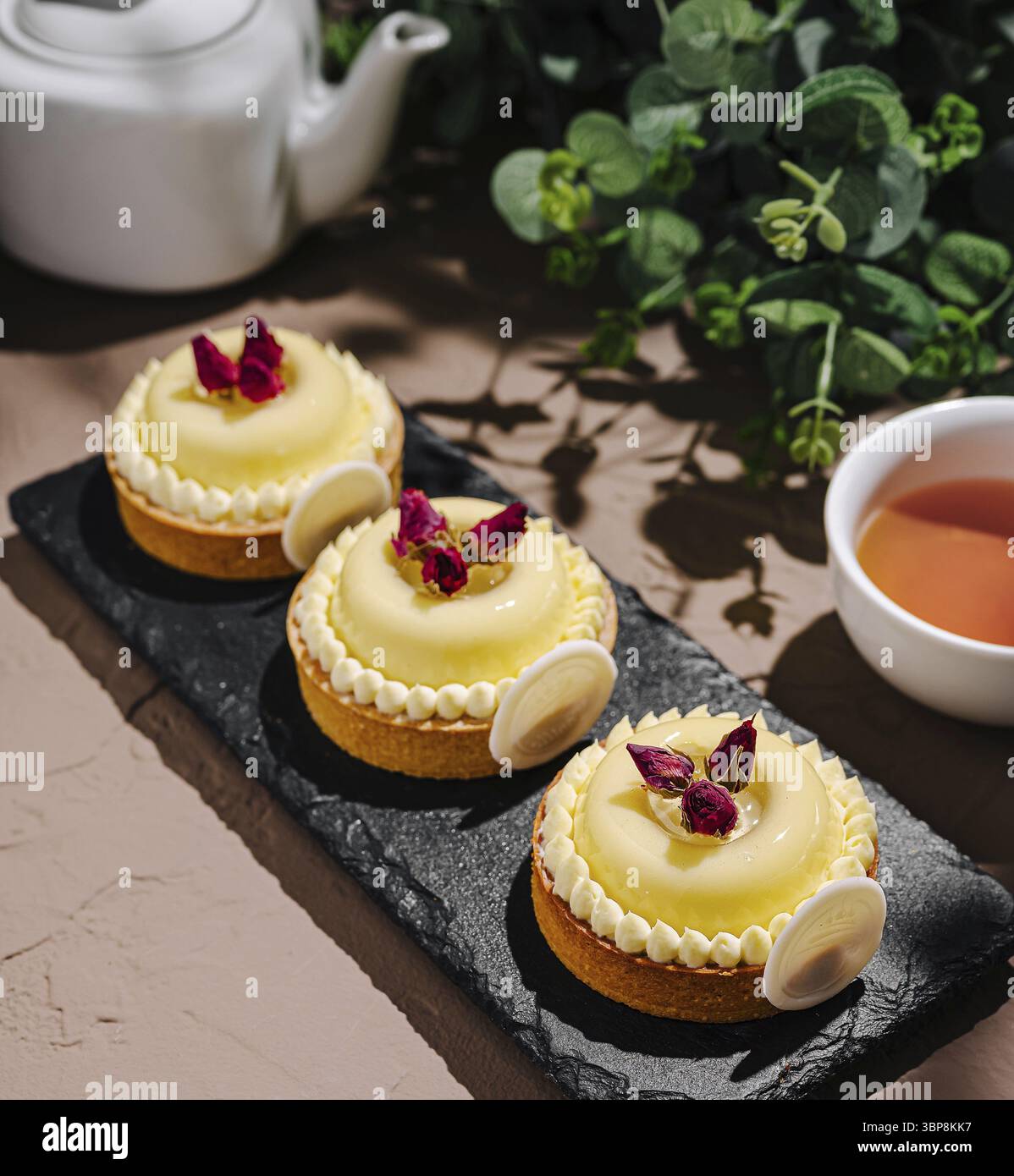 Cibo, tre crostate di limone finemente lavorate sono disposte su un piatto da portata in ardesia. Ogni crostata è ricoperta da petali di rosa e accompagnata da un piccolo fiocco Foto Stock