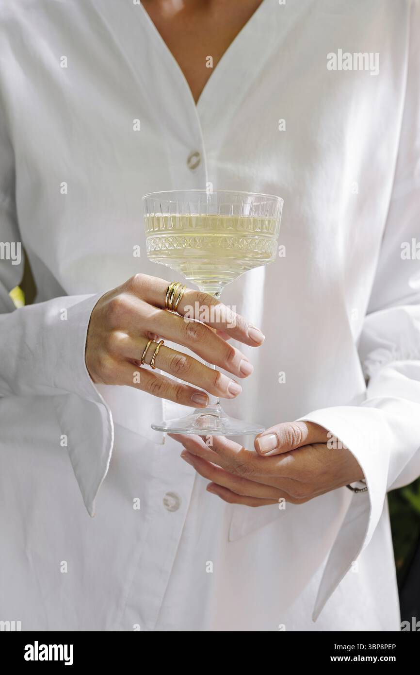Cibo, Una persona tiene un delicato bicchiere di bevanda all'aperto, vestito con una camicia bianca croccante. La luce del sole mette in risalto gli eleganti anelli che adornano le dita Foto Stock