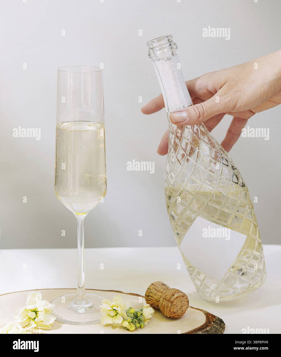 Cibo, Una mano sta versando vino frizzante da una bottiglia in un alto bicchiere di flauto. Il morbido decor floreale e il sughero indicano un'atmosfera festiva, perfetta per Foto Stock