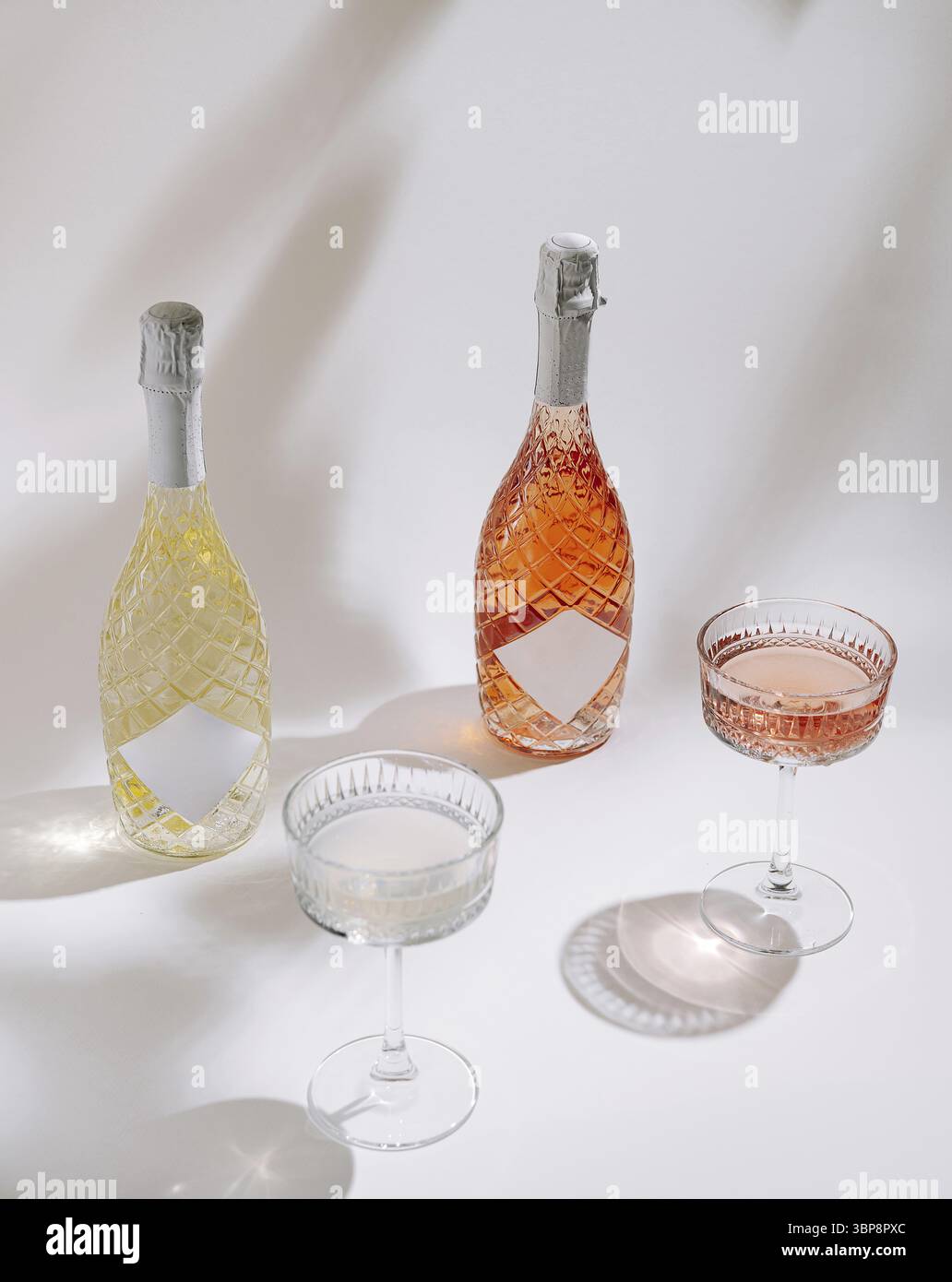 Cibo, due bottiglie di vino frizzante, una gialla e una rosa, siediti accanto a tre eleganti bicchieri pieni di bevande, gettando ombre morbide su un bianco surfato Foto Stock