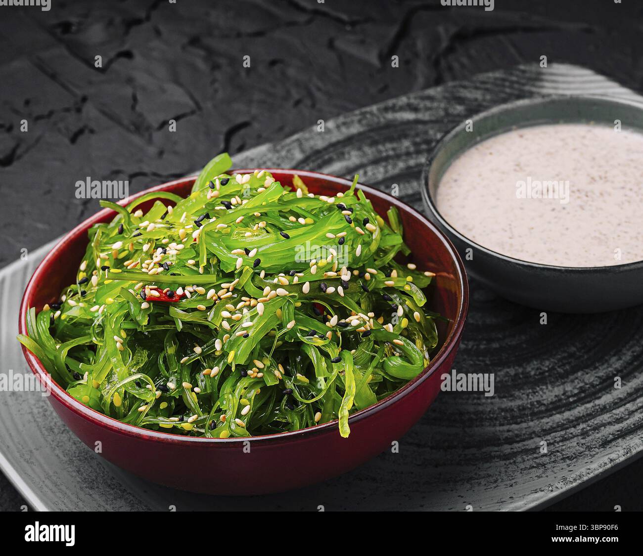 Cibo: Una vivace ciotola di insalata di alghe fresche condita con semi di sesamo si trova su un piatto scuro accanto a una piccola ciotola di condimento cremoso con sesamo. Perfetto per un Foto Stock