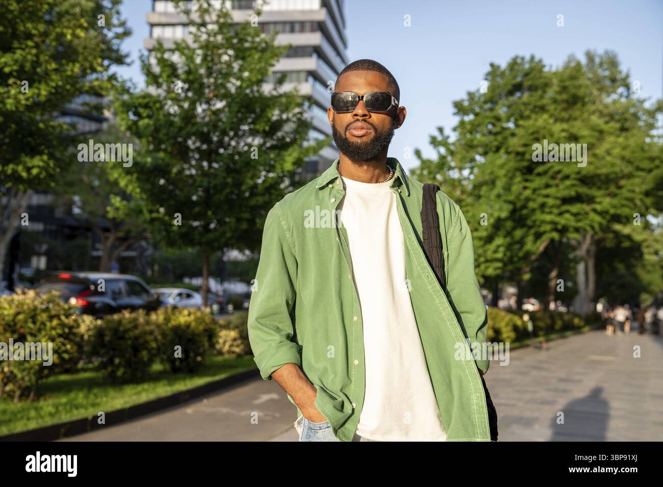 Un uomo nero sorridente cammina lungo il vicolo. Elegante studente afroamericano che vagava per le strade della città con il sole, passeggiando rilassato, rimanendo Foto Stock