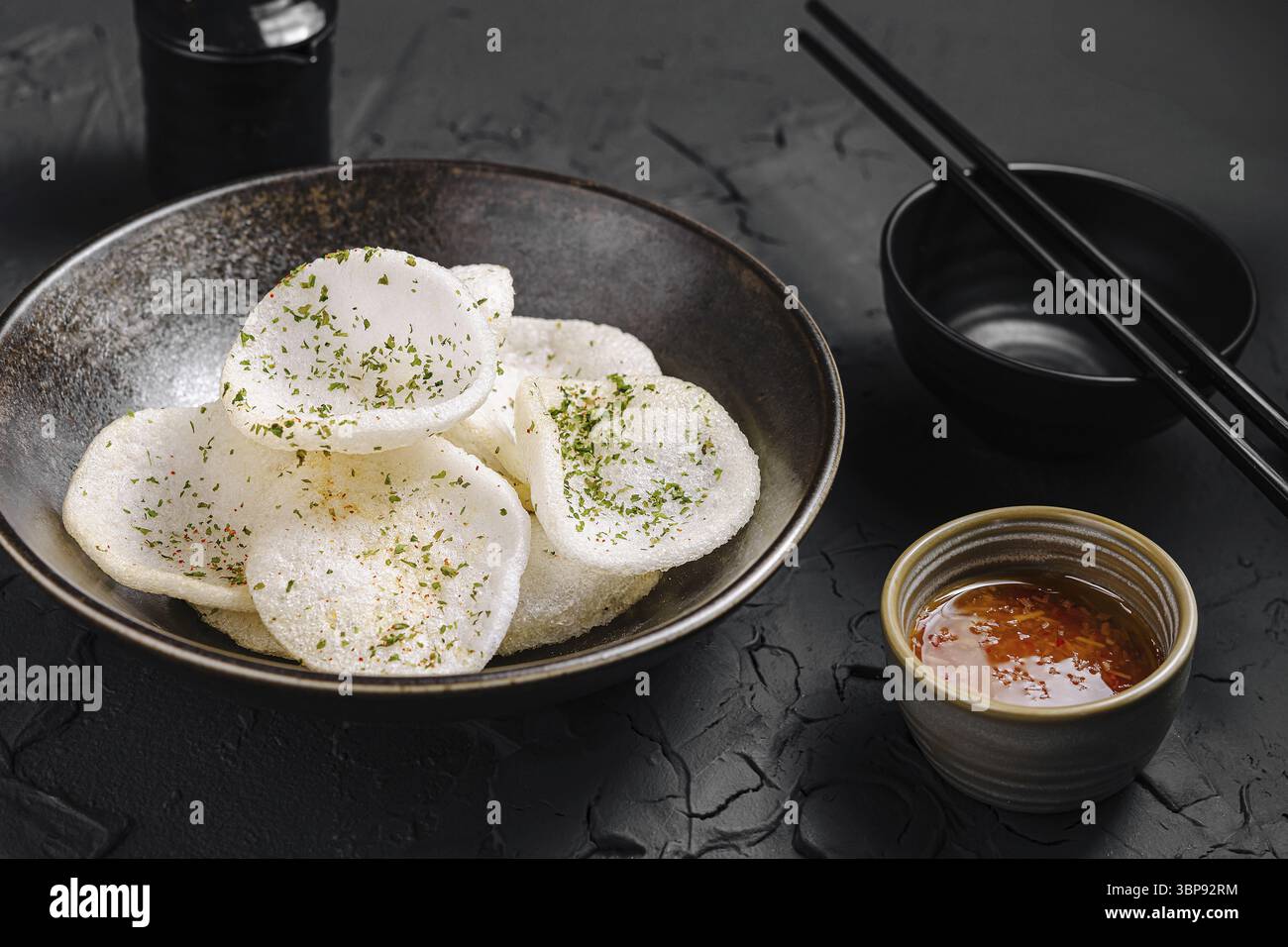 Cibo, gli spuntini bianchi croccanti sono guarniti con erbe in una ciotola scura insieme a un piccolo piatto di salsa piccante e bacchette, creando un'atmosfera accattivante Foto Stock