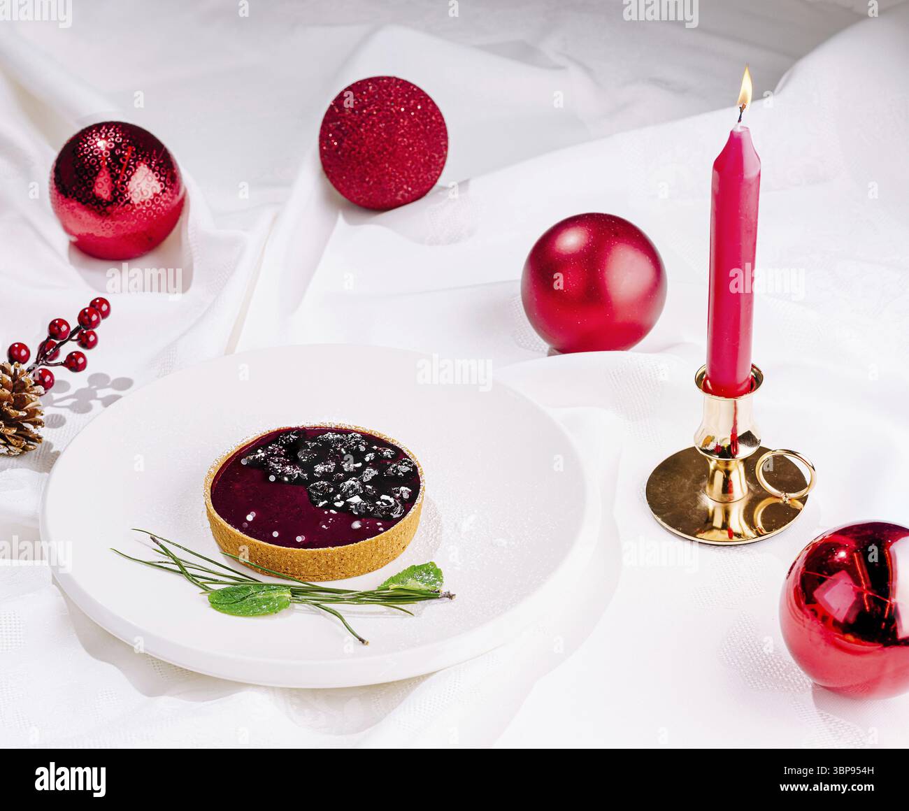 Cibo, Un dessert ben organizzato si trova su un piatto bianco, circondato da festosi ornamenti rossi e una candela. La crostata presenta una crema di frutti di bosco Foto Stock
