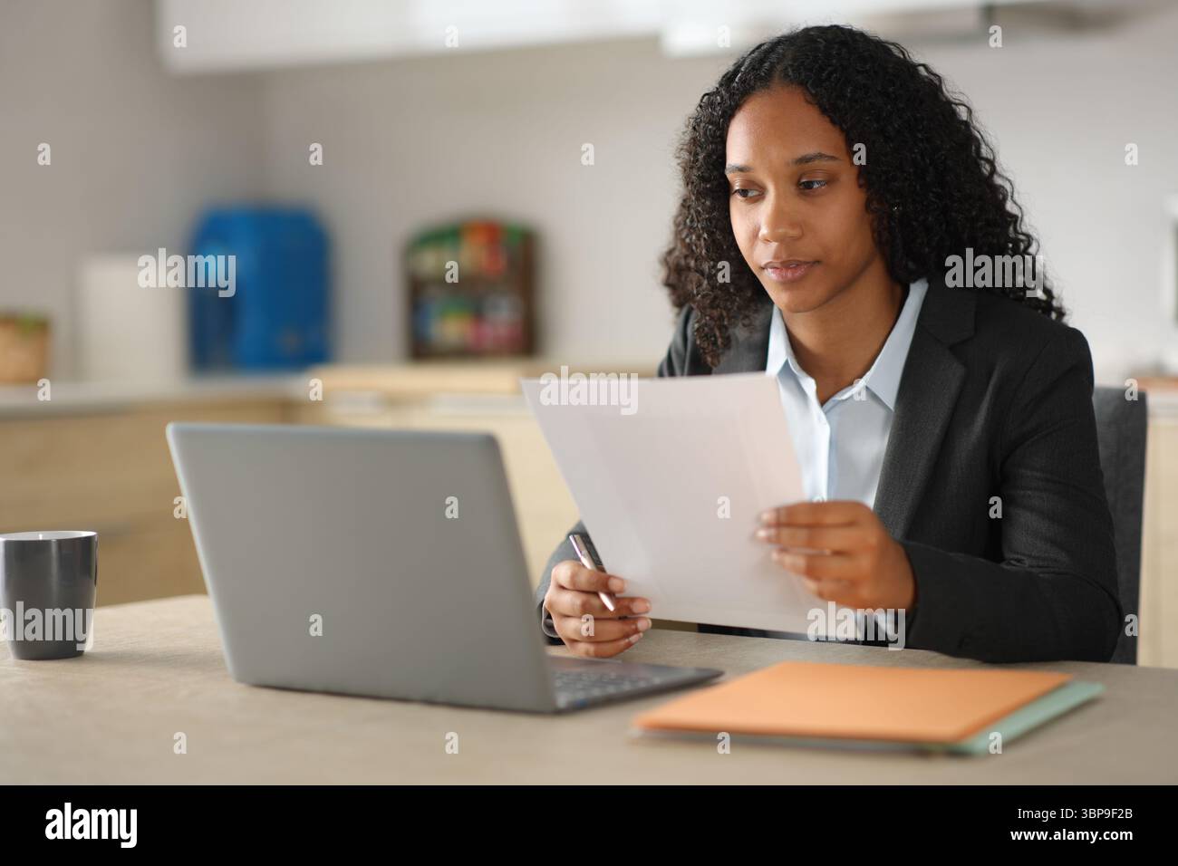 Donna d'affari nera che controlla documenti e laptop in una cucina di casa Foto Stock
