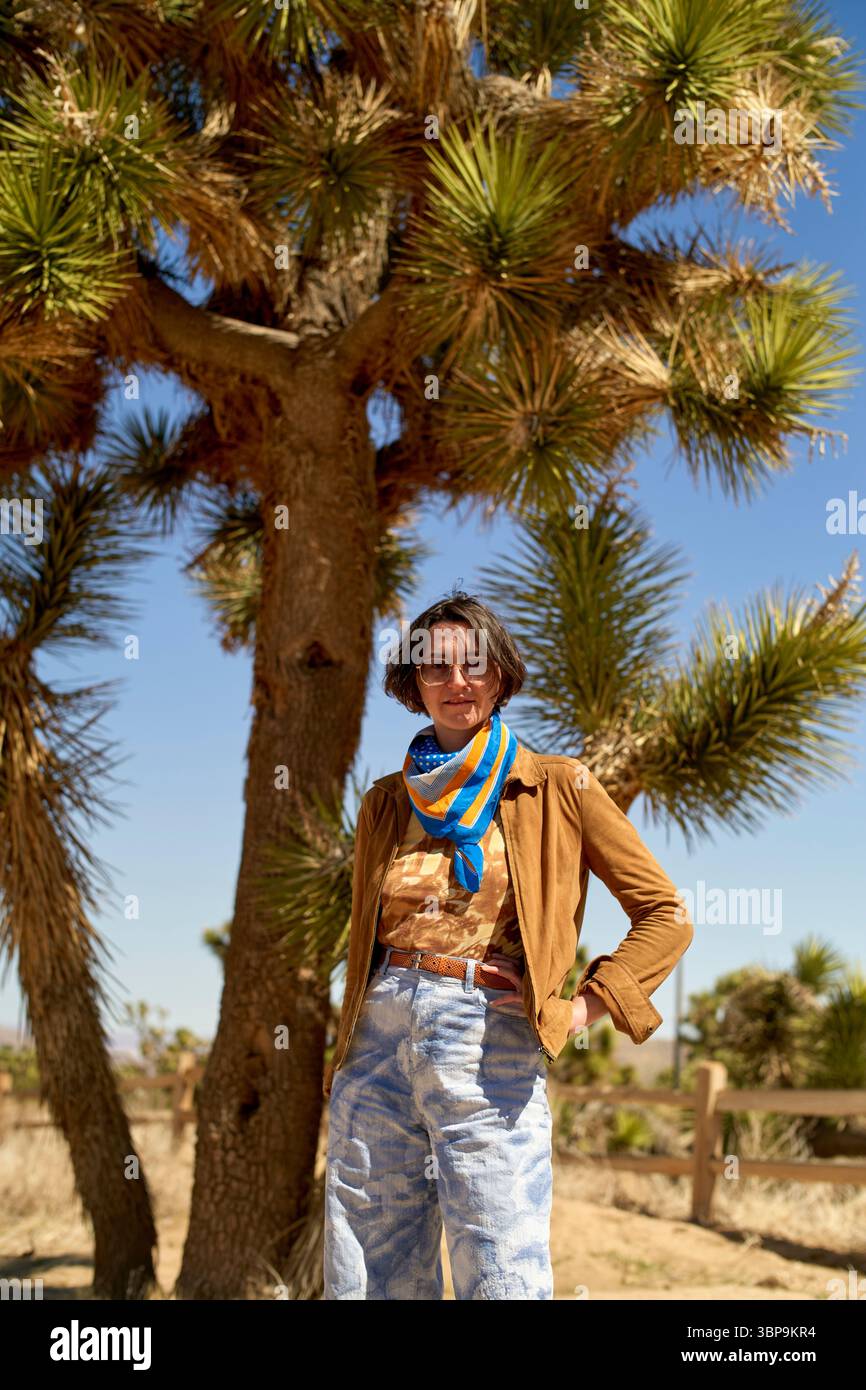 La persona con un abbigliamento casual si trova di fronte a un grande albero in un paesaggio desertico soleggiato. Deserto del Mojave, Stati Uniti Foto Stock