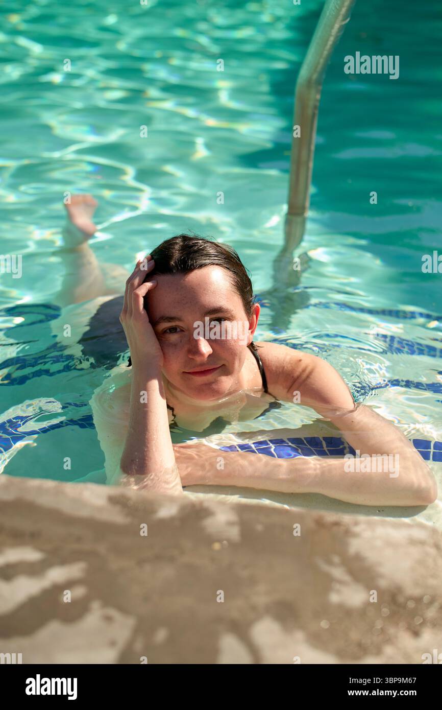 Donna che si rilassa in piscina, appoggiata su un gradino, con acqua cristallina intorno a lei. Palm Springs, Stati Uniti Foto Stock