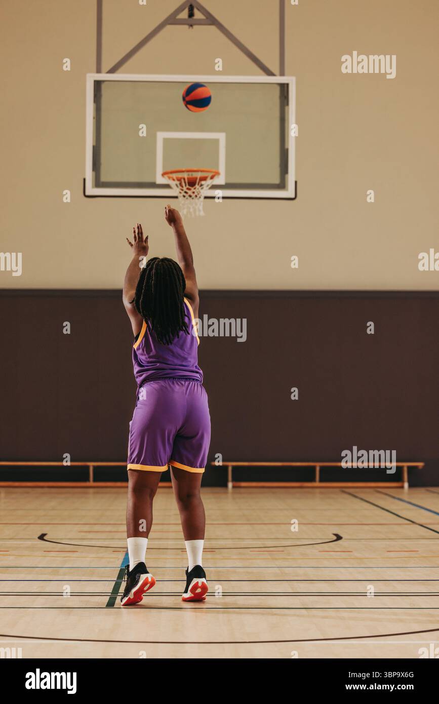 Un giovane che indossa una divisa da basket viola lancia un pallacanestro nel cerchio durante la pratica, mostrando abilità atletiche e concentrazione in un moderno Foto Stock