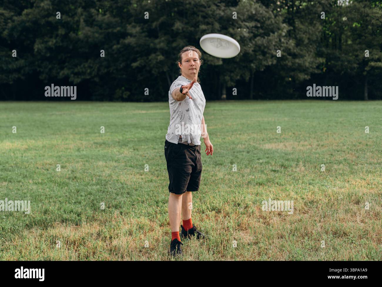Uomo che lancia un disco frisbee con estensione del braccio diretta verso la fotocamera attraverso l'erba verde in piena luce naturale. Foto Stock