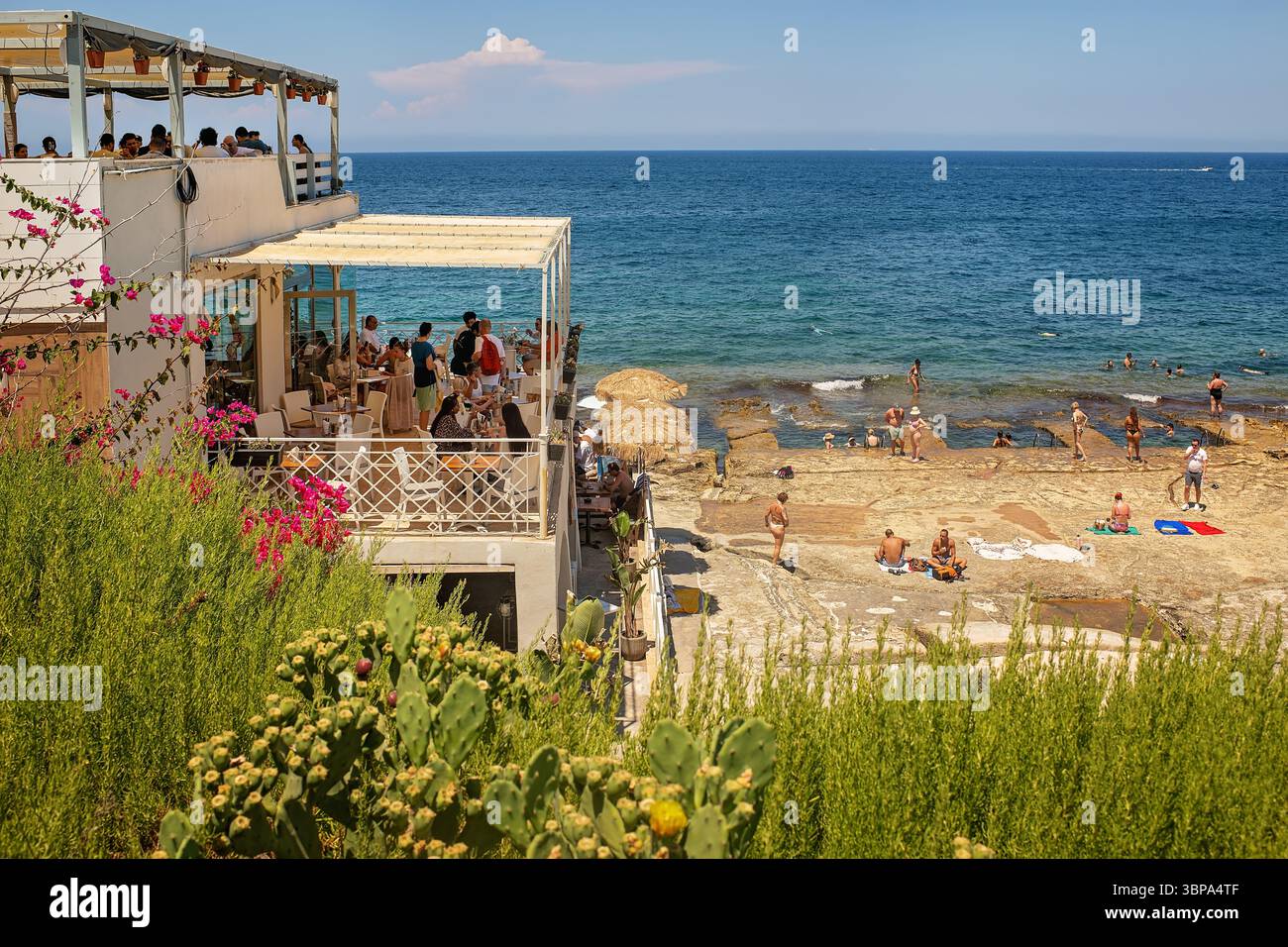 Sliema, Malta - 06/07/2025: Una vista attraverso una lussureggiante vegetazione fino ad un affollato caffè a due piani e alla spiaggia della città con piscine di roccia naturale e turisti Foto Stock