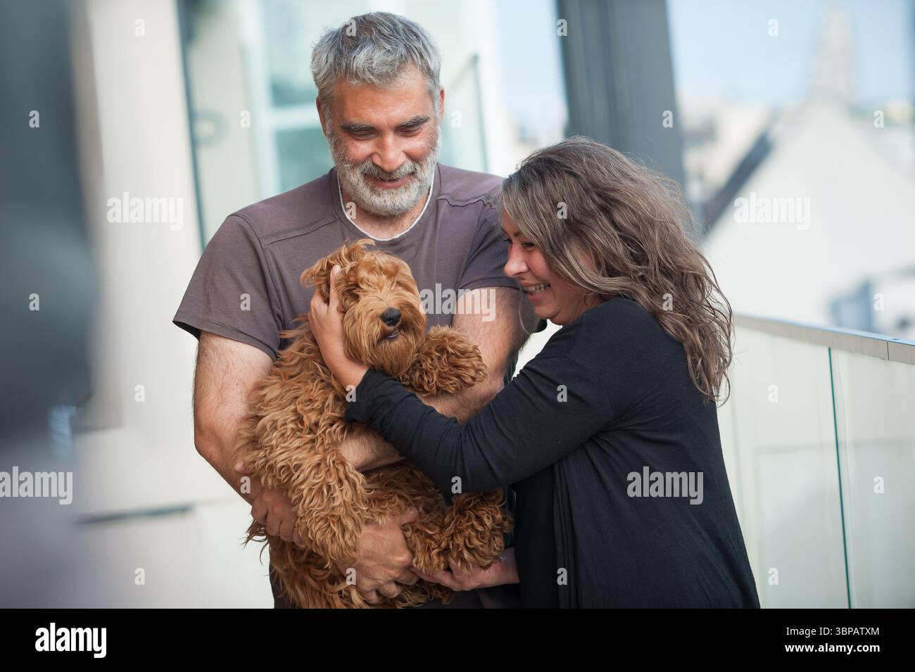 Un uomo e una donna sorridenti stanno insieme tenendo il loro adorabile cane irlandese Terrier. Sono su un balcone che guarda il loro animale domestico con affetto. Vista sulla città A. Foto Stock