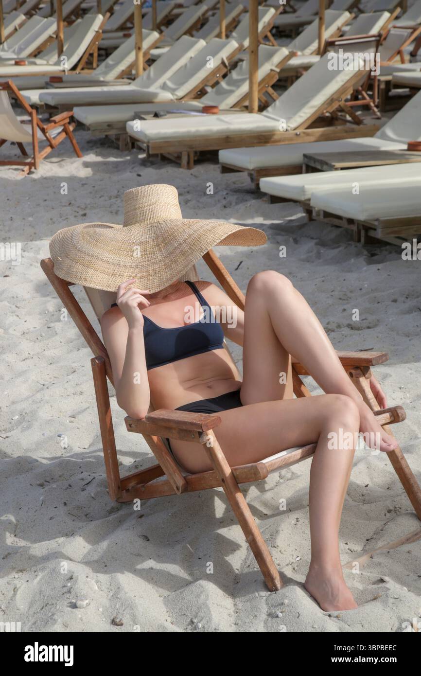 Rilassata giovane donna che prende il sole sulla sedia da spiaggia in legno nascondendo il viso sotto un cappello di paglia largo, godendosi una giornata idilliaca in spiaggia. Vacanza in stile tropicale Foto Stock