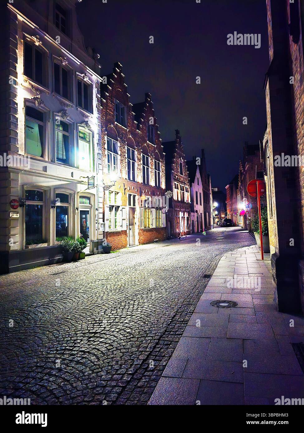 Tranquilla strada acciottolata di notte a Bruges, Belgio, fiancheggiata da una storica architettura fiamminga e da una soffusa illuminazione stradale. - Immagine stock catturata con smartphone