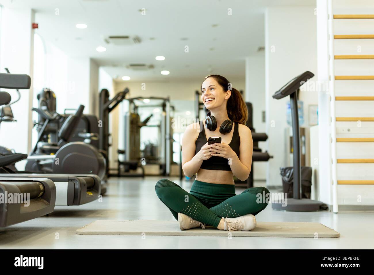 Sorridendo entusiasticamente tenendo le cuffie, una giovane donna gode di un momento di beatitudine in un vivace centro fitness. Foto Stock