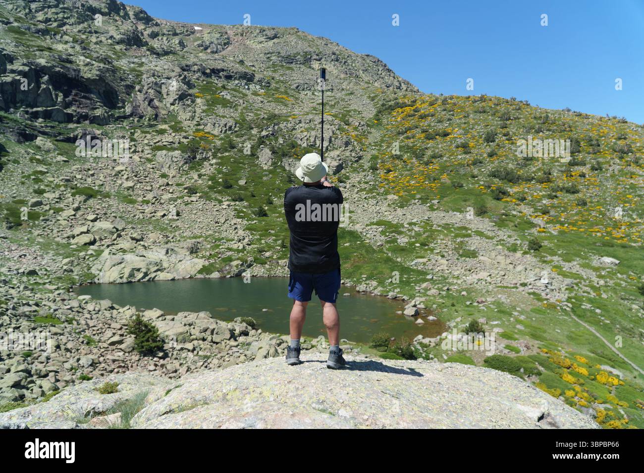 Un uomo è in piedi su una roccia che guarda un lago. Indossa una camicia nera e pantaloncini blu Foto Stock