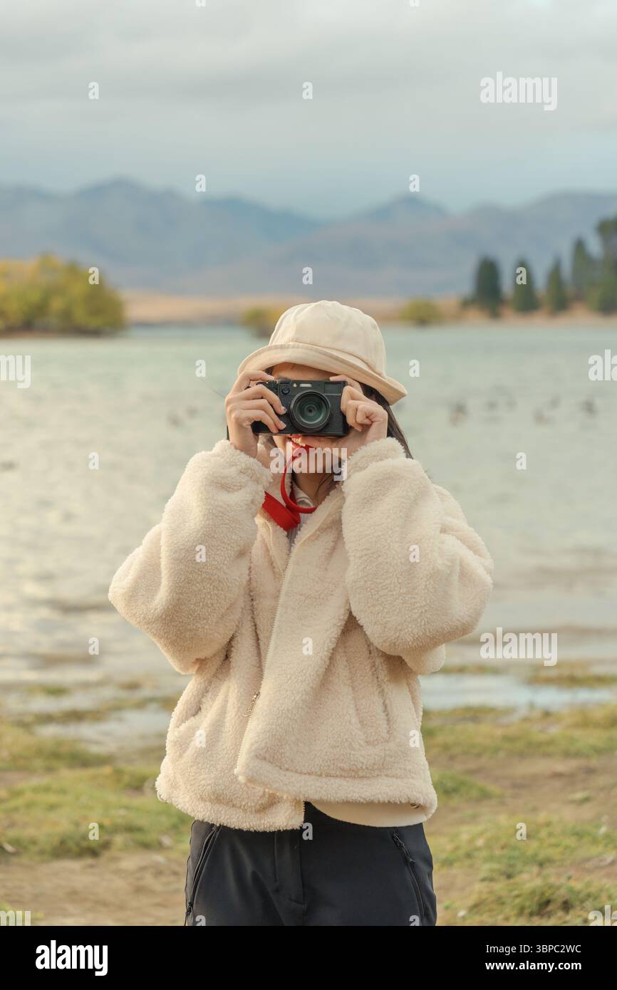 Giovane turista asiatico in un'accogliente giacca, cappello che si gode scattando foto con una macchina fotografica retrò sul lago Tekapo in autunno in nuova Zelanda Foto Stock