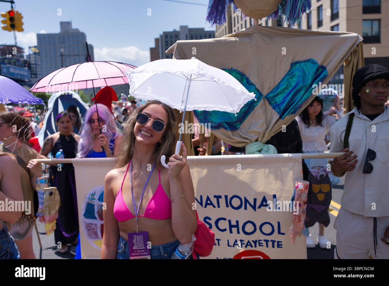 Una giovane donna con gli occhiali da sole tiene un ombrellone bianco in una vivace sfilata. Indossa un bikini rosa e un cordone. Dietro di lei, un grande display galleggiante Foto Stock