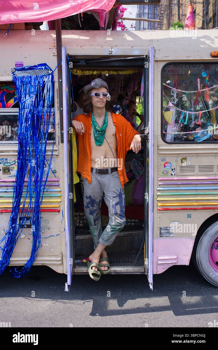 Una persona con un colore abbronzato, che indossa una camicia arancione, pantaloni grigi con disegni dipinti, occhiali da sole e una bandana turchese, si erge sulla porta Foto Stock