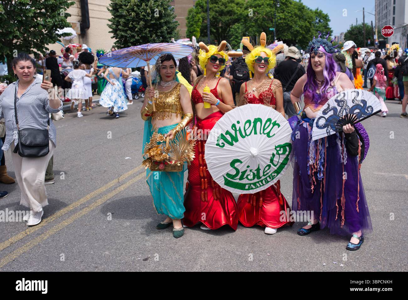 Quattro donne in costumi elaborati, tra cui code di sirena e panini hot dog, partecipano a una sfilata di strada. Hanno ombrelloni e ventilatori, con un par Foto Stock