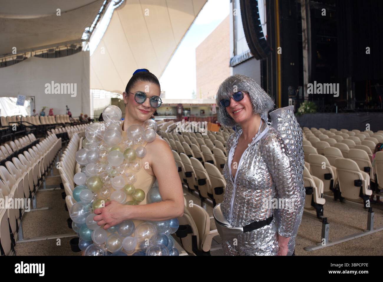 Due donne con un abbigliamento insolito si trovano in un anfiteatro all'aperto. Uno indossa un abito decorato con bolle di plastica trasparente, l'altro un salto argentato con paillettes Foto Stock